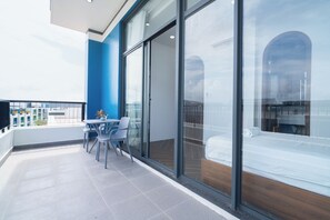 Een modern balkon met een tafel en stoelen, uitzicht op de stad door grote glazen deuren, en een blauwe accentmuur.