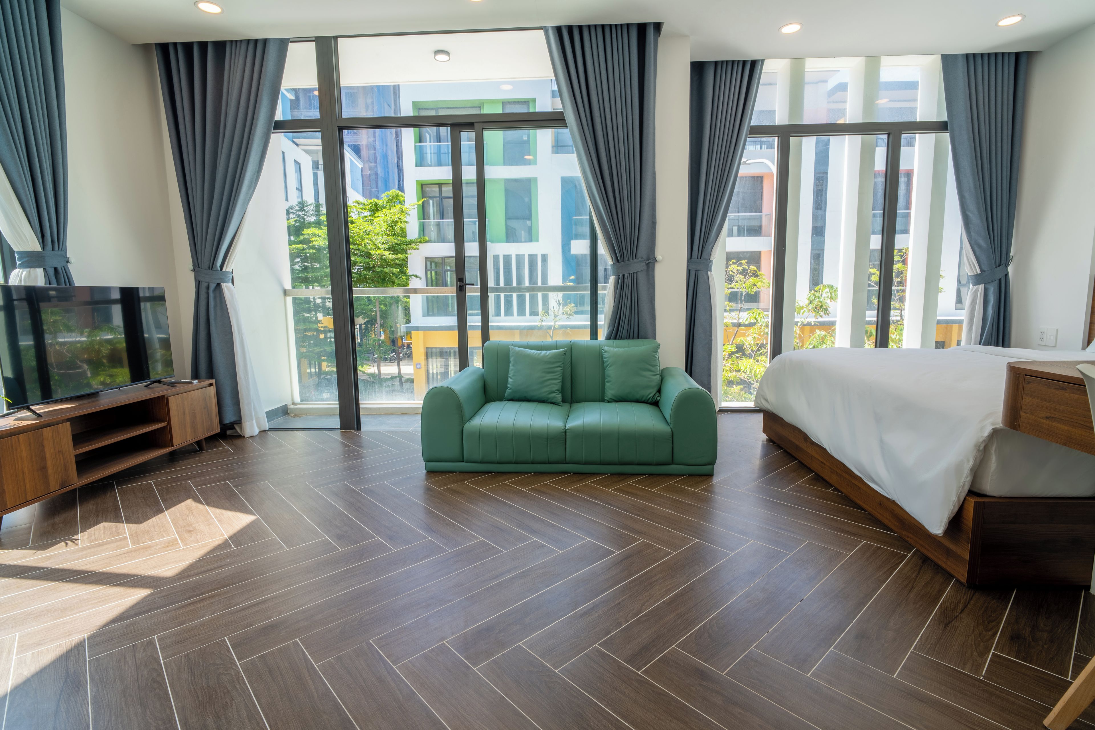 Una habitación de hotel moderna con un sofá verde, una cama, un televisor y ventanas grandes con cortinas.