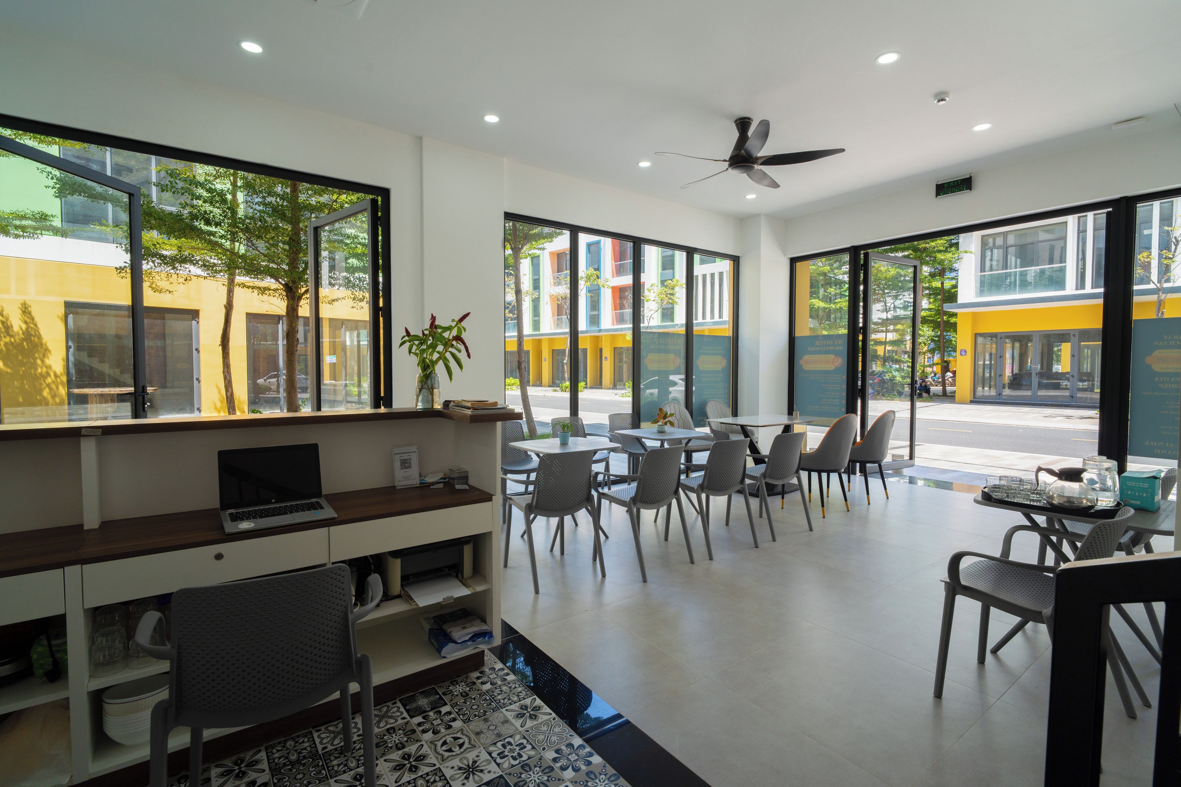 A modern cafe with a laptop on a desk, chairs around a table, and large windows offering a view of the street.