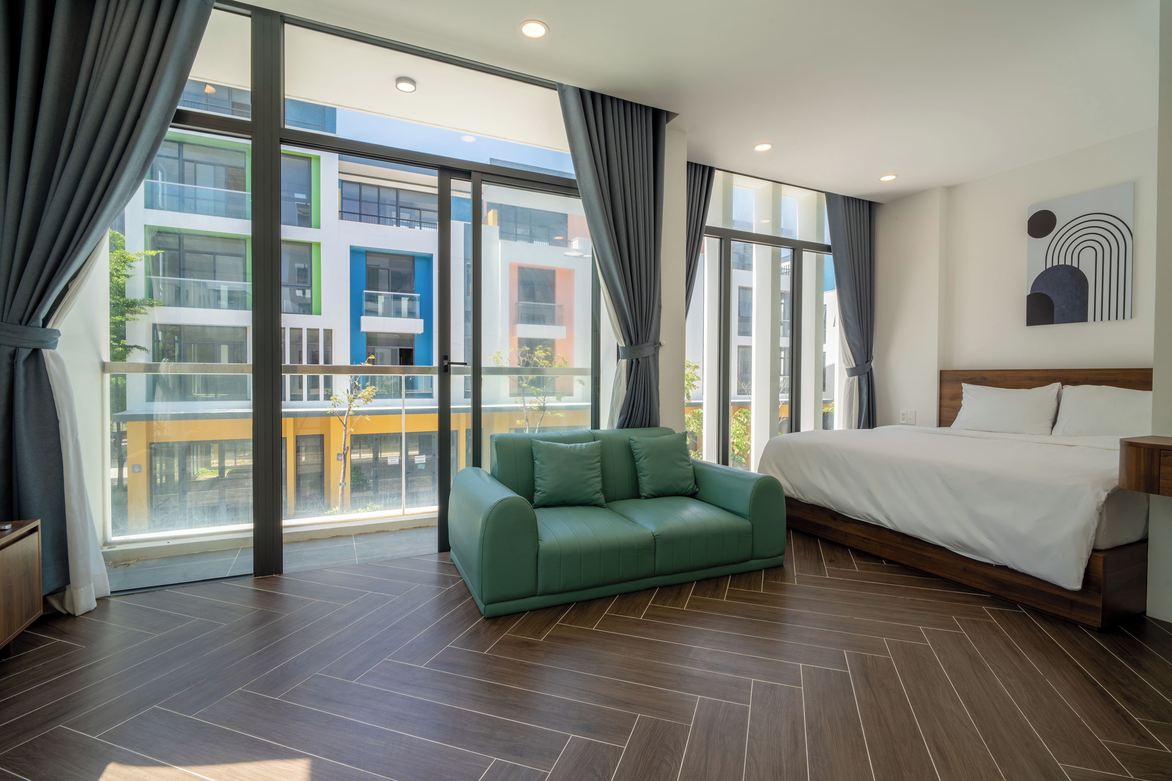 Une chambre d’hôtel moderne avec un grand lit, un canapé vert et un balcon offrant une vue sur un bâtiment aux couleurs vives.