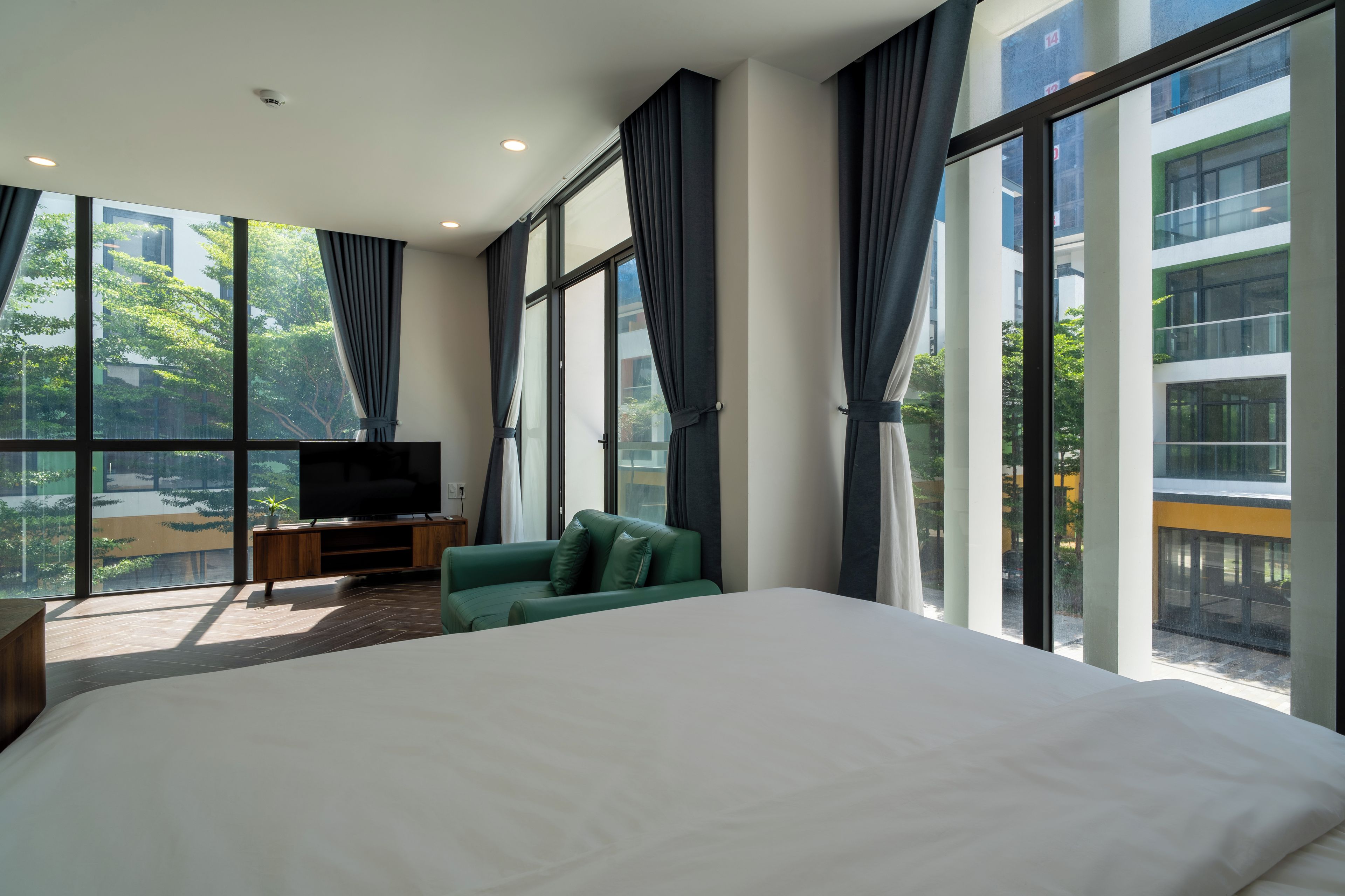 A modern hotel room with a large bed, a green sofa, a TV on a wooden stand, and large windows offering a view of the city.