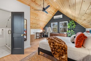 Ein Schlafzimmer mit schrägen Wänden, einem Bett mit brauner Decke, einem Schreibtisch mit Stuhl und einem Fenster mit Blick auf Bäume.