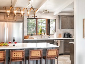 A modern kitchen with a large island, stainless steel appliances, and a view of trees through large windows. - Private Estate | Sport Court | Hot Tubs | Saunas (Asheville)