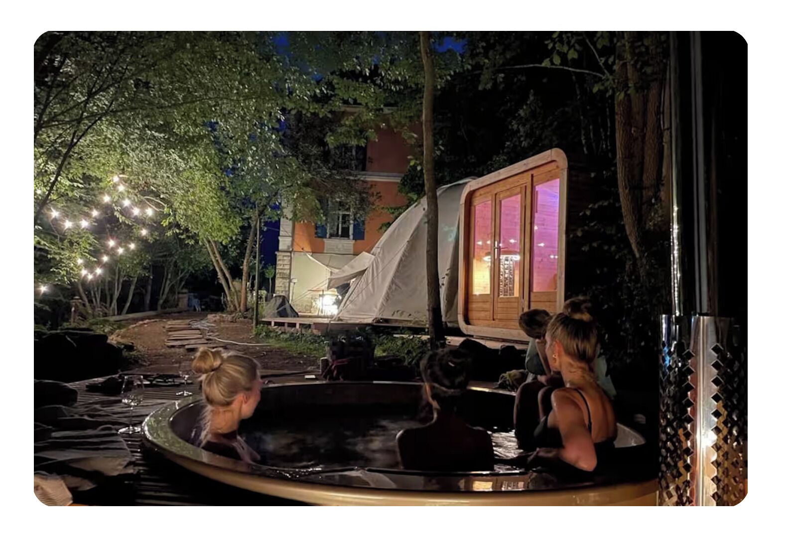 A hot tub with a wooden deck, surrounded by trees and a tent.