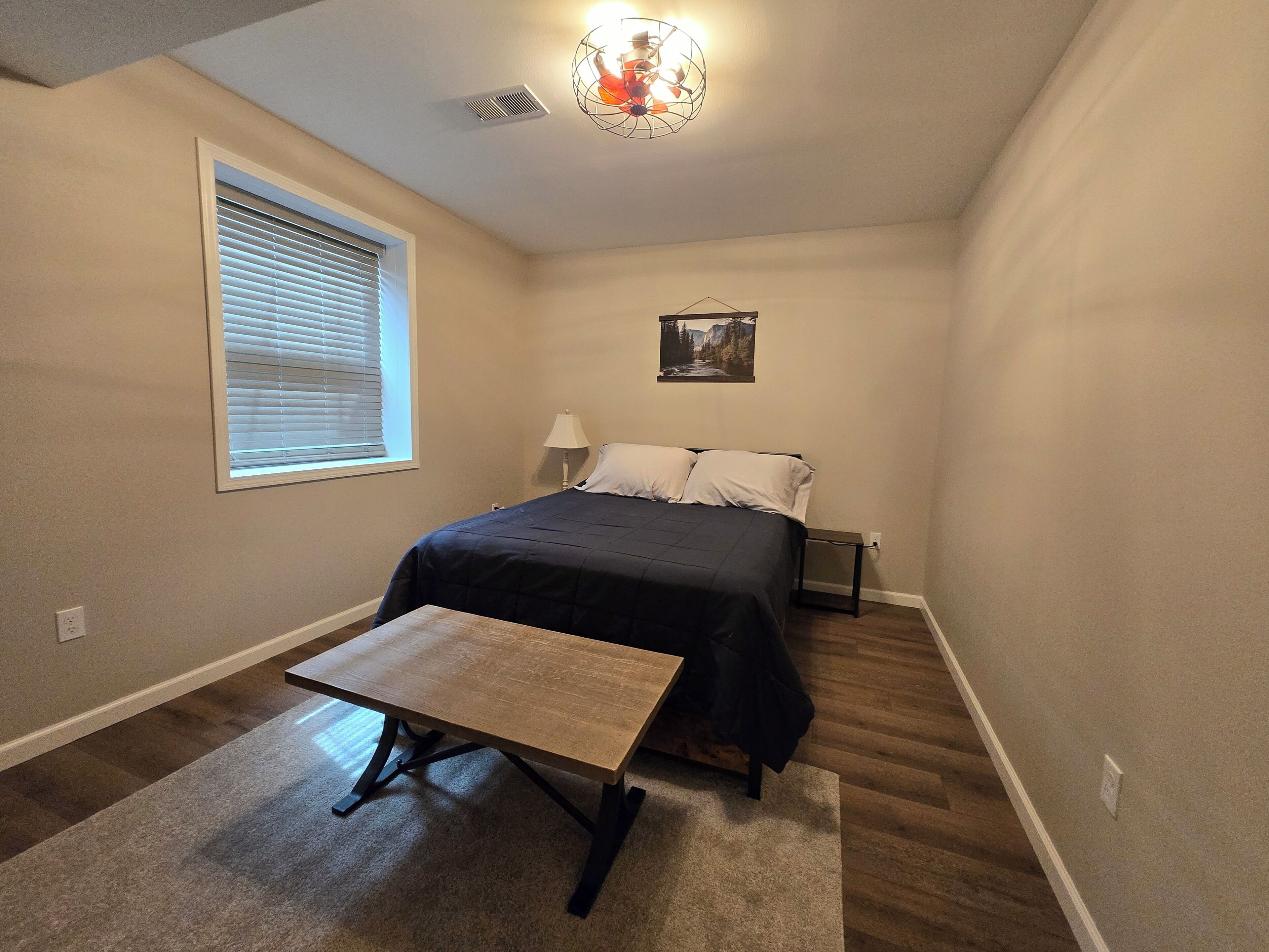 A bedroom with a bed, a bedside table, a lamp, a window with blinds, a ceiling light fixture, and a wall-mounted picture.