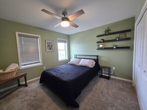 A bedroom with a bed, a ceiling fan, a window with blinds, a bedside table, and wall shelves with decorative items.