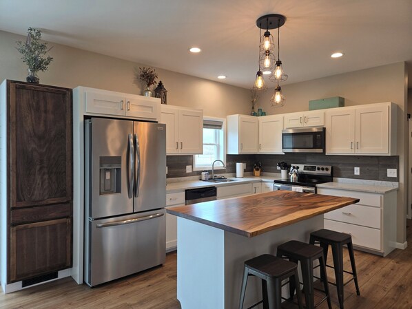 A modern kitchen with a stainless steel refrigerator, white cabinets, a wooden island, bar stools, and pendant lighting.