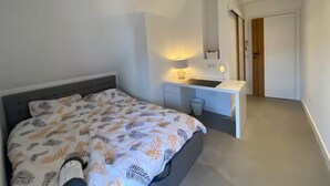 A bedroom with a bed, a bedside table with a lamp, a ceiling fan, and a door leading to another room.