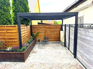 Un espacio cubierto con un patio de madera, un pérgola de metal y dos sillas.