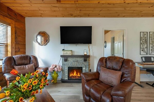 A living room with a fireplace, leather chairs, a TV, and a vase of flowers.