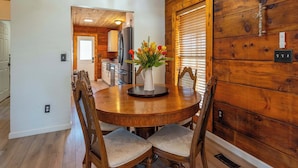 A wooden interior with a dining table, chairs, a vase with flowers, and a kitchen in the background.