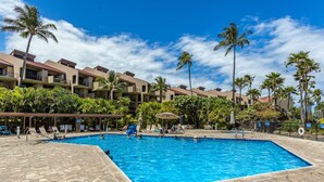 A swimming pool with lounge chairs, palm trees, and a building with balconies. - Kamaole Sands Condo | Ground Floor, 2 Bath, Pool, Hot Tub, AC, Walk to Beach (Kihei)