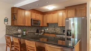 A kitchen with wooden cabinets, a stainless steel refrigerator, a microwave, and a tiled backsplash. - Kamaole Sands Condo | Ground Floor, 2 Bath, Pool, Hot Tub, AC, Walk to Beach (Kihei)