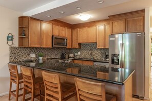 A kitchen with wooden cabinets, a stainless steel refrigerator, a microwave, and a tiled backsplash. - Kamaole Sands Condo | Ground Floor, 2 Bath, Pool, Hot Tub, AC, Walk to Beach (Kihei)