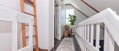 A narrow hallway with a wooden ladder, a white railing, and a rug leading to a door.