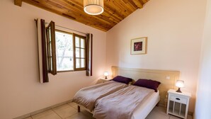 Een slaapkamer met een houten plafond, een bed met twee kussens, een nachtkastje met een lamp en een raam met gordijnen.