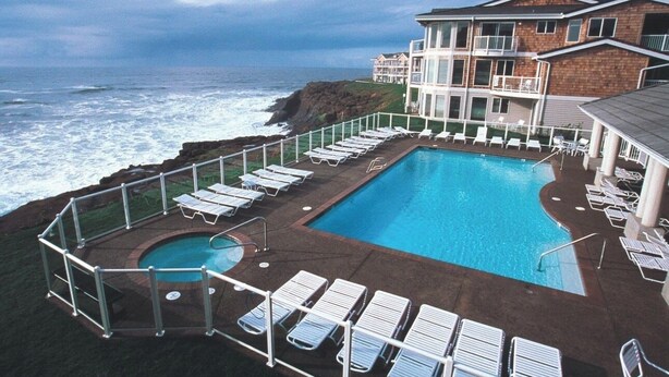 A pool with lounge chairs, a building with balconies, and a view of the ocean. - CRASHING WAVES AND WHALES! (Depoe Bay)