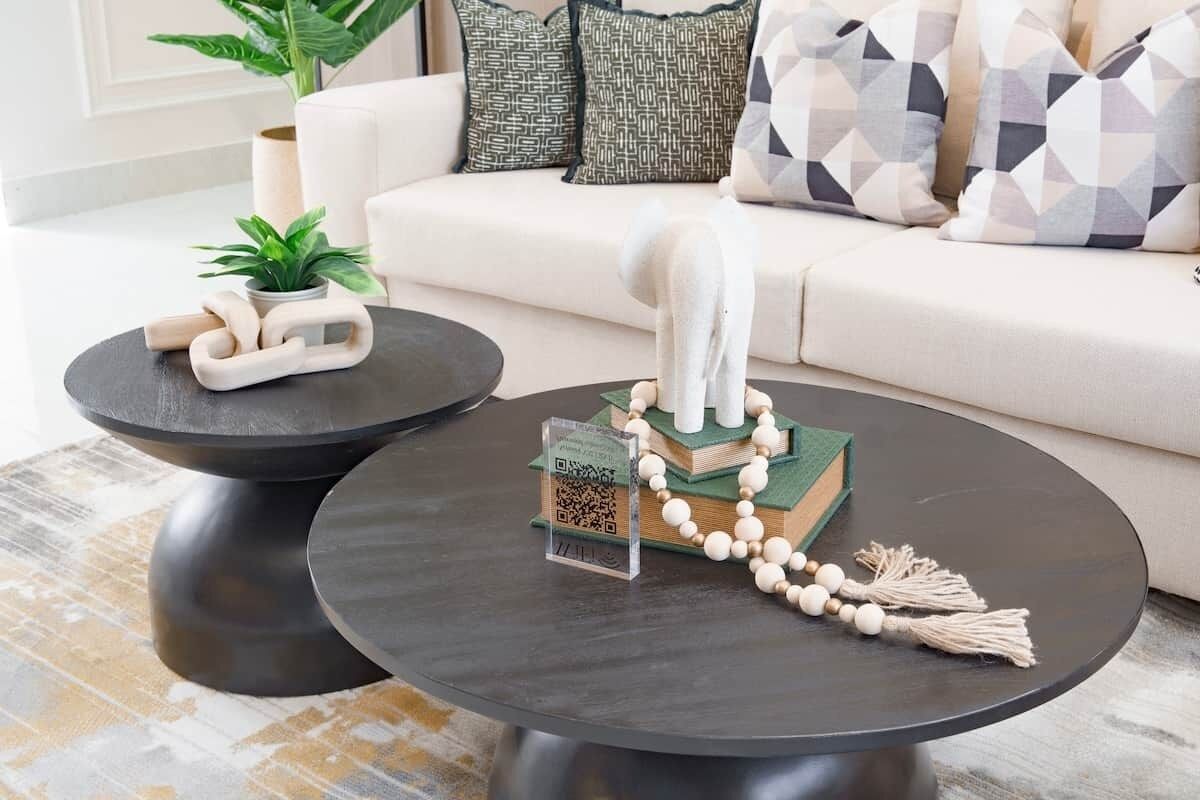 Un salon moderne avec un canapé, deux tables basses, des coussins décoratifs, une petite plante et un livre agrémenté d’un collier de perles.