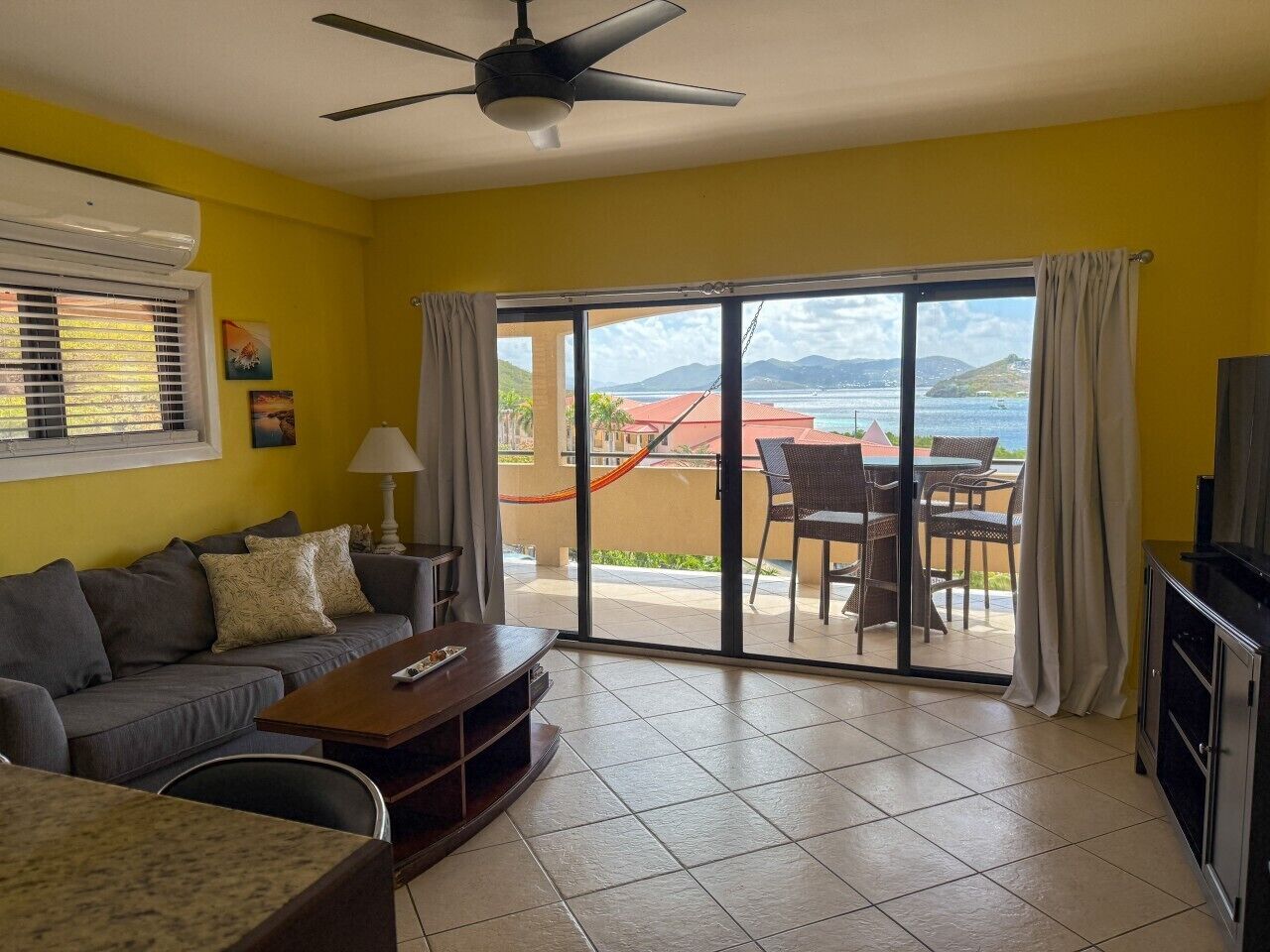 A living room with a sofa, coffee table, and TV stand. There is a sliding glass door leading to an outdoor area with a view of the sea and mountains.
