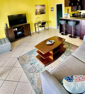 A living room with a TV, a wooden coffee table, a sofa, and a kitchen area with bar stools. - Red Hook Retreat with Stunning Ocean Views (Red Hook)