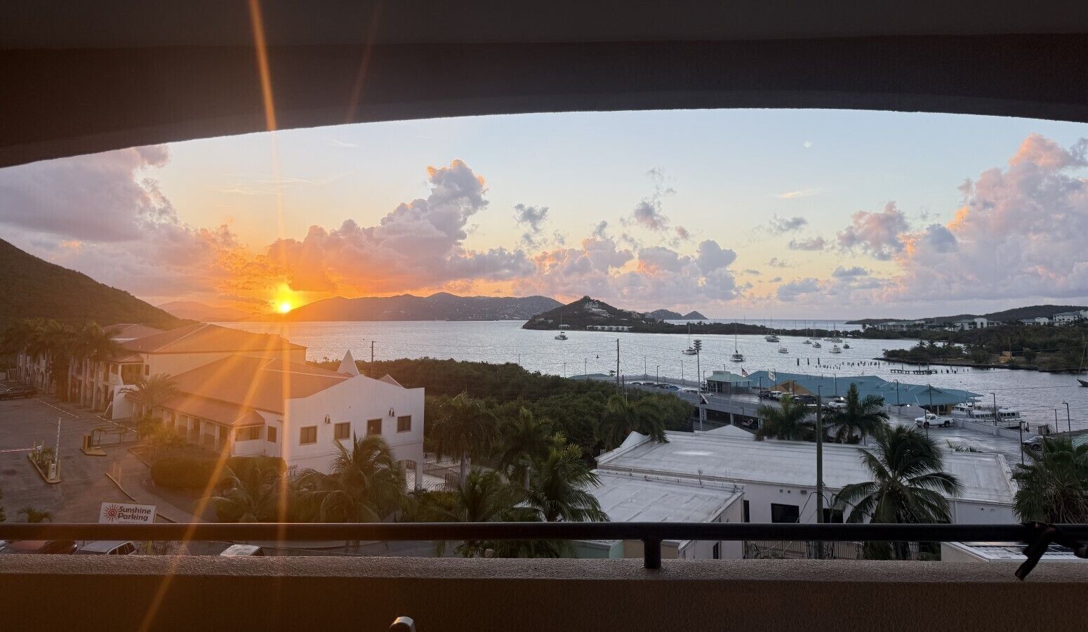 Une vue sur le coucher de soleil depuis un balcon donnant sur une marina, avec des bateaux, un parking et des bâtiments.