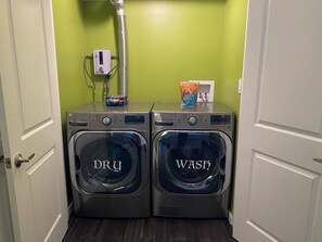 A laundry area with two front-loading washing machines labeled 'DRY' and 'WASH,' a water heater, and a Tide detergent box. - Spacious 2BR Retreat • Balcony Views + Parking (Red Hook)