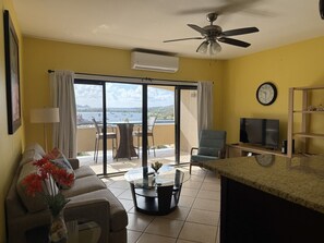 Ein Wohnzimmer mit einer Couch, einem Couchtisch, einem Fernseher und einem Balkon mit Meerblick.