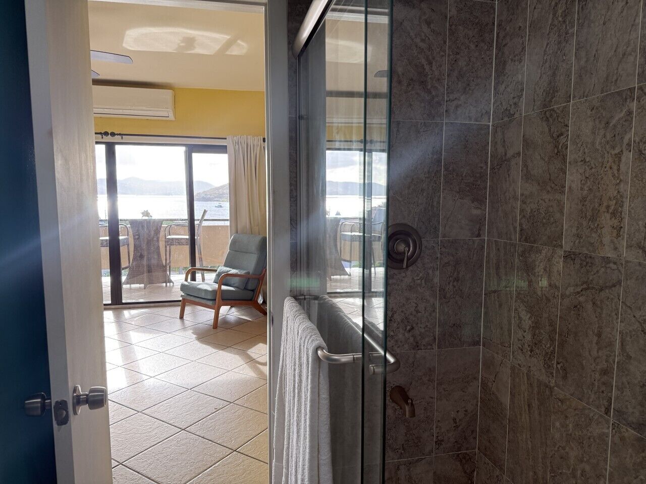 A bathroom with a glass shower door, a towel hanging on the door, and a view of an outdoor balcony with a chair and table.