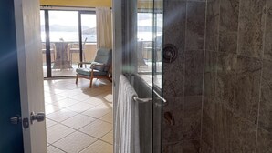 A bathroom with a glass shower door, a towel hanging on the door, and a view of an outdoor balcony with a chair and table. - Walk to Ferry • Red Hook 1BR Condo w/ Stunning Balcony Views + Free Parking (Red Hook)