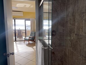 A bathroom with a glass shower door, a towel hanging on the door, and a view of an outdoor balcony with a chair and table. - Walk to Ferry • Red Hook 1BR Condo w/ Stunning Balcony Views + Free Parking (Red Hook)