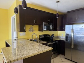 A kitchen with granite countertops, stainless steel appliances, dark wooden cabinets, and a yellow accent wall. - Walk to Ferry • Red Hook 1BR Condo w/ Stunning Balcony Views + Free Parking (Red Hook)