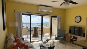 A living room with a sofa, a coffee table, a TV, and a balcony with a view of the sea. - Walk to Ferry • Red Hook 1BR Condo w/ Stunning Balcony Views + Free Parking (Red Hook)