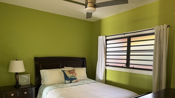 A bedroom with a bed, a nightstand with a lamp, a window with curtains, and a ceiling fan. - Walk to Ferry • Red Hook 1BR Condo w/ Stunning Balcony Views + Free Parking (Red Hook)