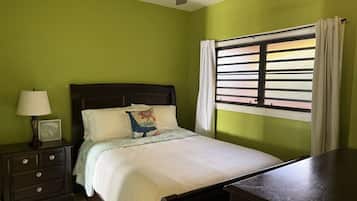 A bedroom with a bed, a nightstand with a lamp, a window with curtains, and a ceiling fan.