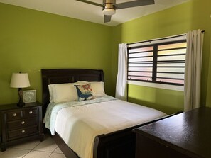A bedroom with a bed, a nightstand with a lamp, a window with curtains, and a ceiling fan. - Walk to Ferry • Red Hook 1BR Condo w/ Stunning Balcony Views + Free Parking (Red Hook)