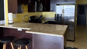 A kitchen with dark wooden cabinets, a stainless steel refrigerator, and a granite countertop. There are three hanging pendant lights and a black trash bin. - Walk to Ferry • Red Hook 1BR Condo w/ Stunning Balcony Views + Free Parking (Red Hook)