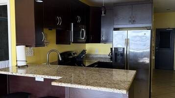 A kitchen with dark wooden cabinets, a stainless steel refrigerator, and a granite countertop. There are three hanging pendant lights and a black trash bin.