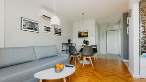 A modern living room with a grey sofa, a round coffee table with oranges, a dining area with chairs and a table, and a hallway leading to a door. - Shortstaypoland Mokotowska (Warsaw)