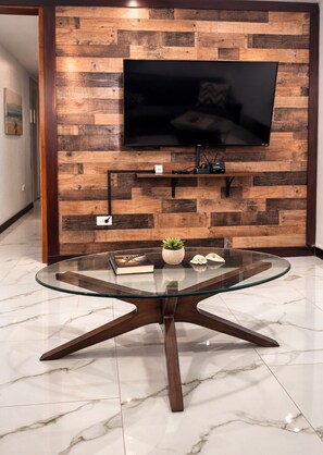 Ein modernes Wohnzimmer mit einem Glastisch, einem Wand-Fernseher und einer Holzwand.