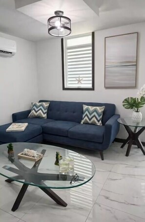 Ein modernes Wohnzimmer mit einem blauen Sofa, einem Glastisch, einer Deckenleuchte und einem gerahmten Bild an der Wand.