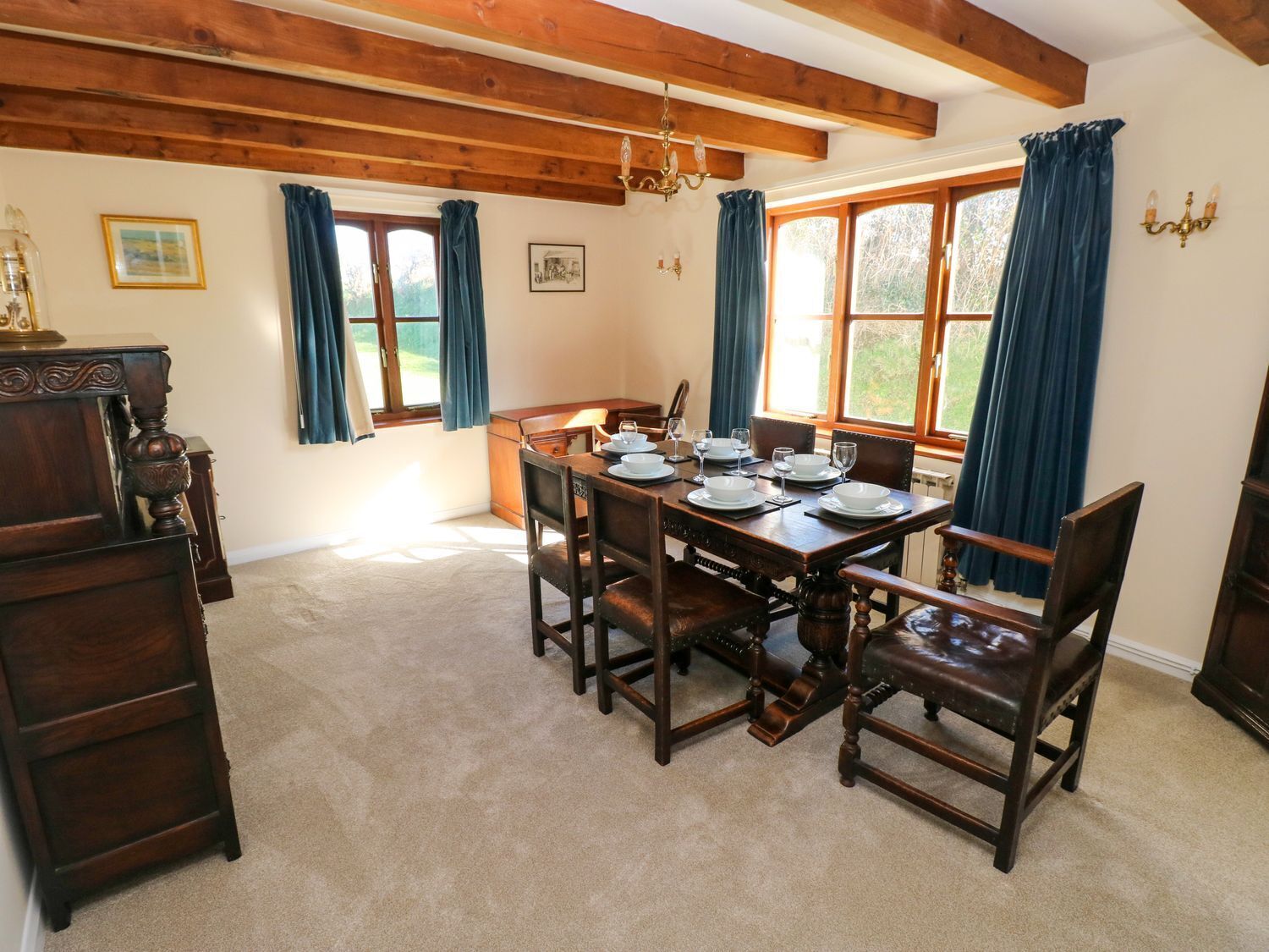 A dining room with a wooden table set for four, surrounded by chairs, with a chandelier above and a window with curtains.