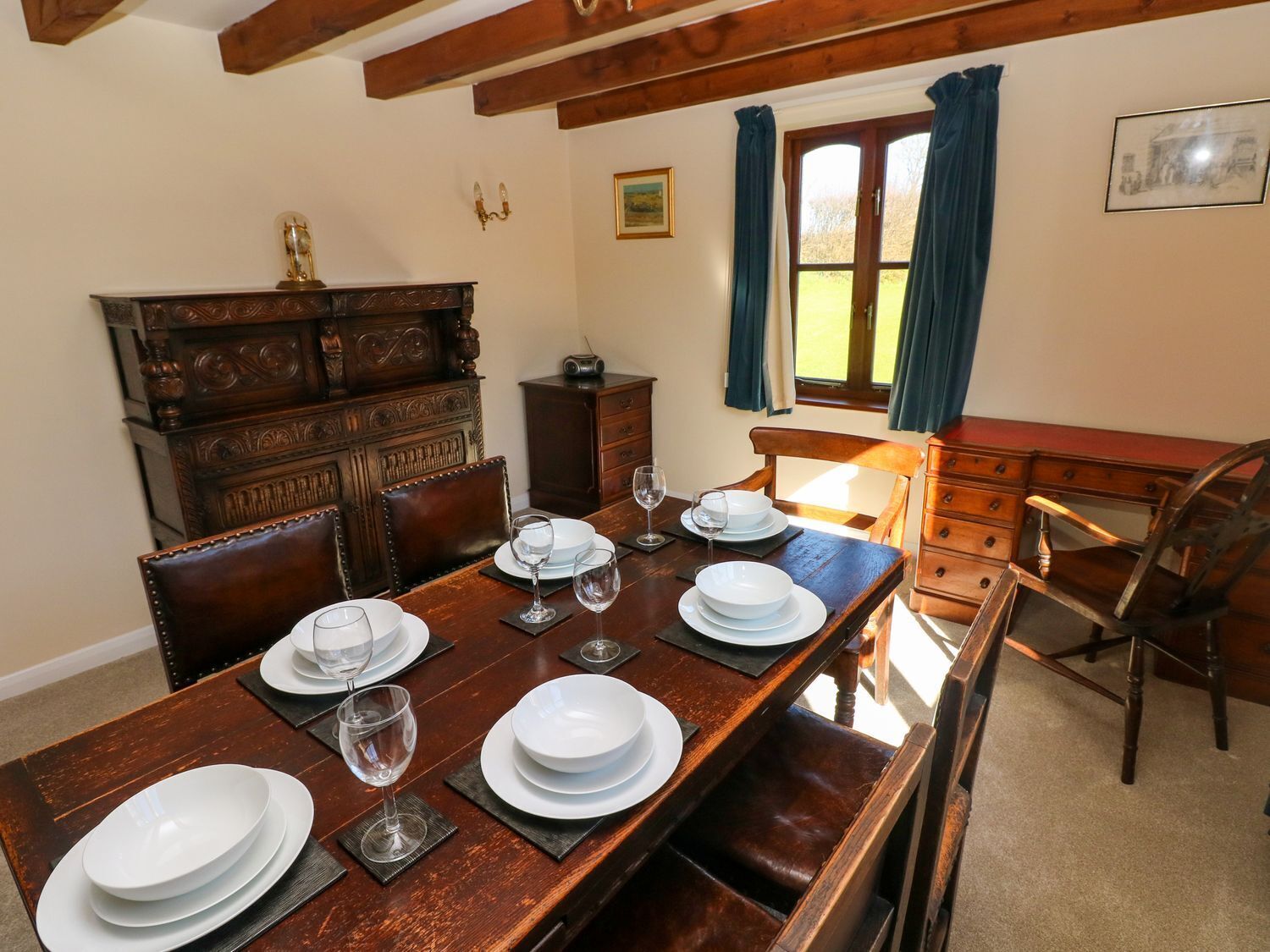 A dining room with a wooden table set for four, a wooden cabinet, a desk, and a window with curtains.