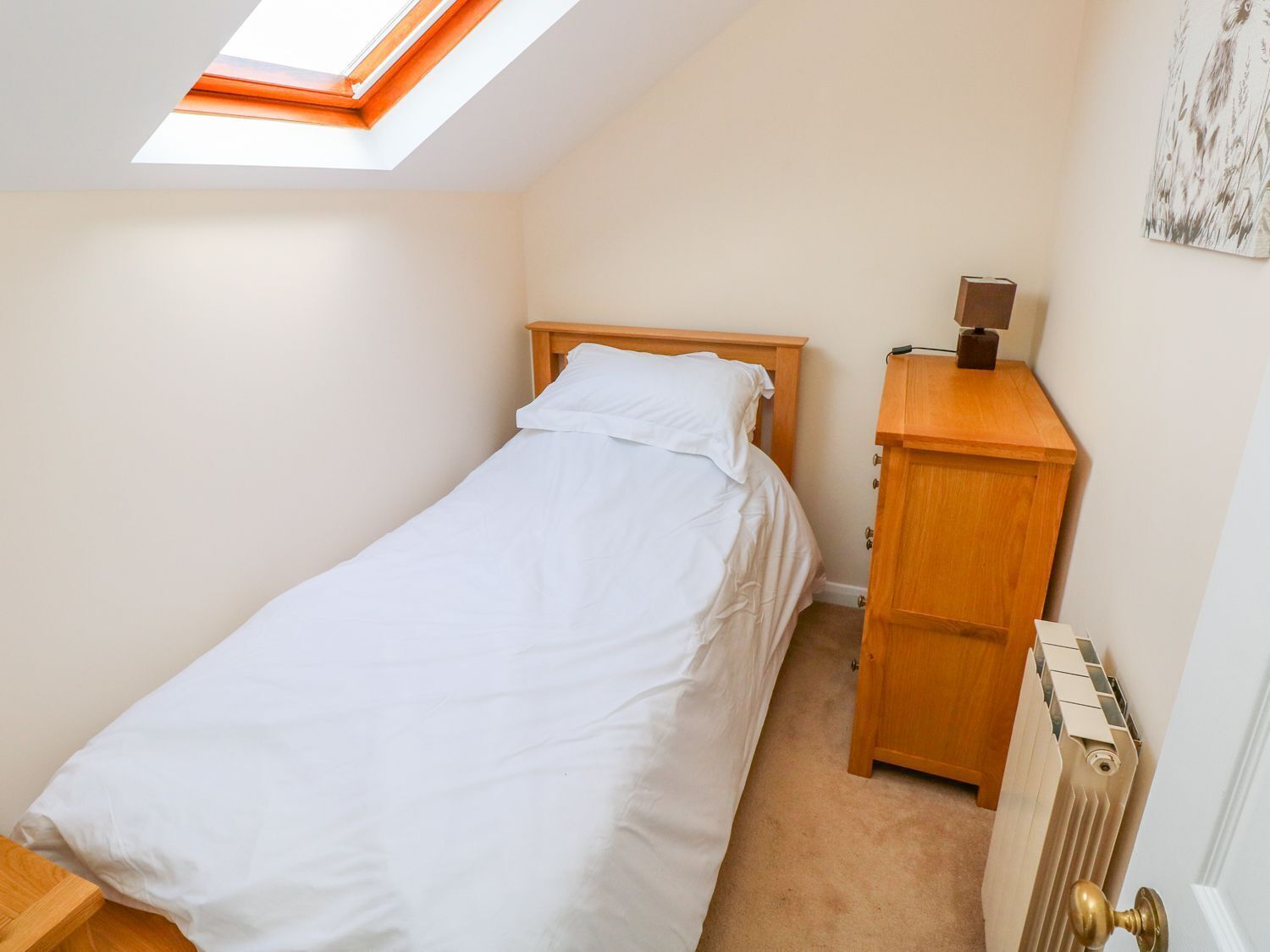 A small, single-bed room with a skylight, a wooden bedside table, and a radiator.