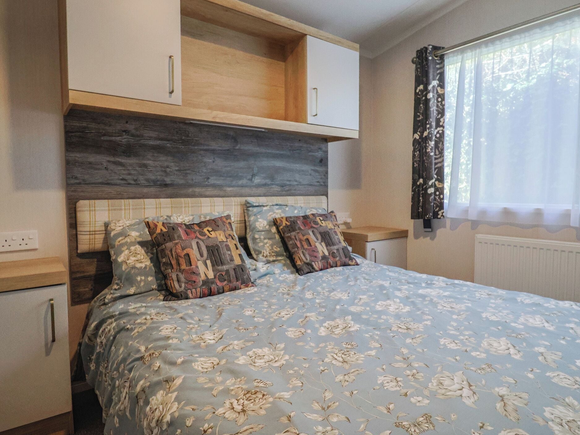 Un dormitorio con cama, dos almohadas, cabecera de madera, ventana con cortinas y un radiador.