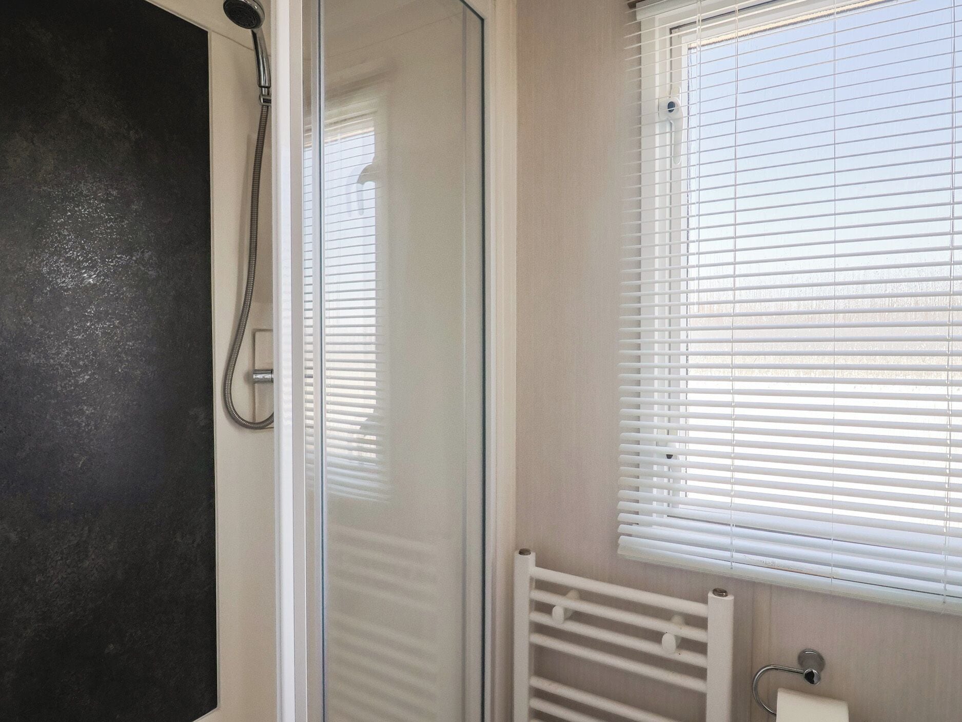 A shower with a glass door, a window with blinds, and a white radiator.