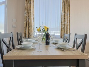 Une table à manger dressée avec des assiettes, des verres et une bouteille, entourée de chaises, avec une fenêtre et des rideaux en arrière-plan.
