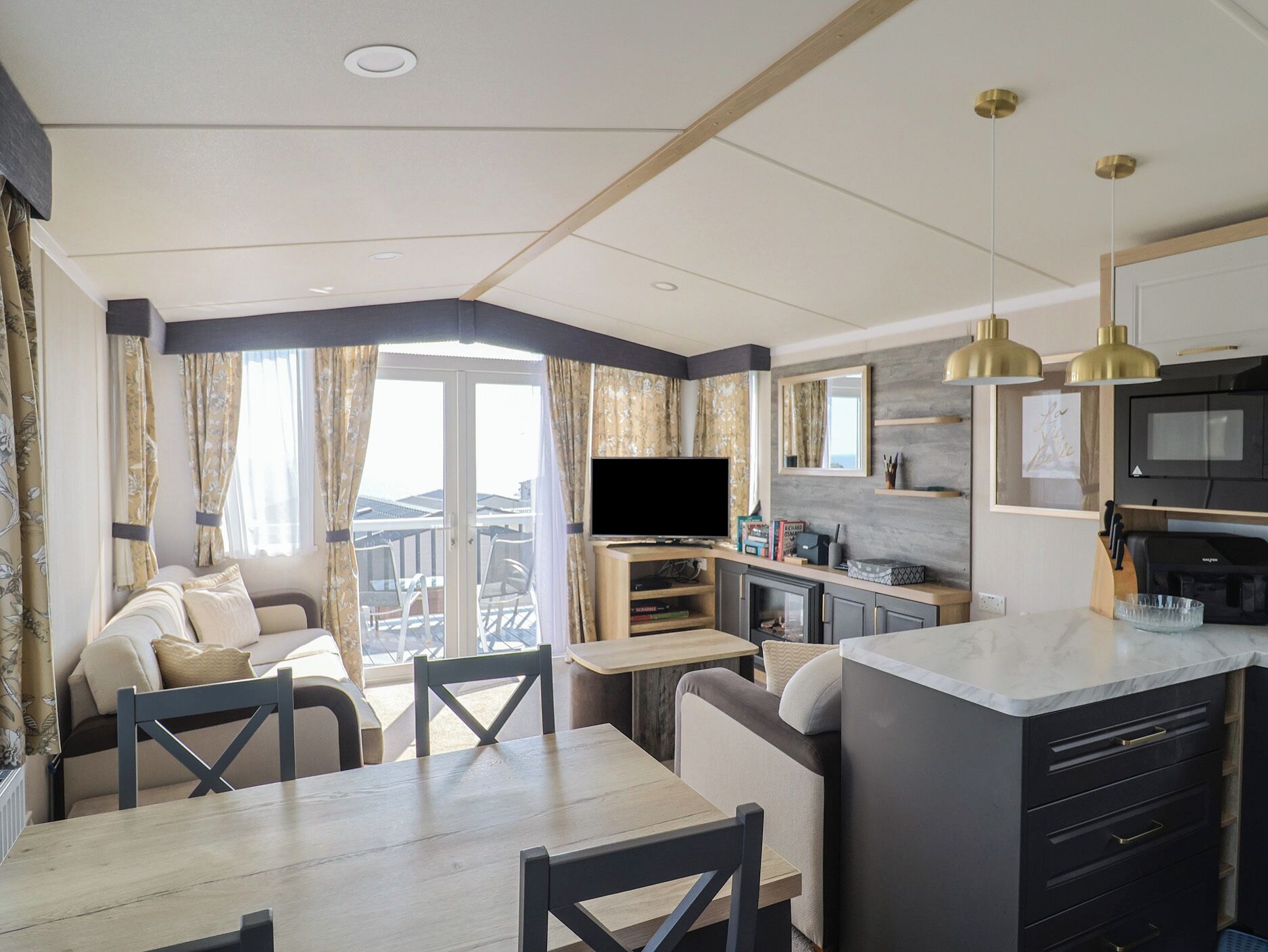 Una sala moderna con sofá, mesa de comedor y sillas, un área de cocina con televisor, y un balcón con cortinas.