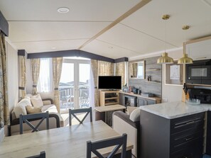 A modern living room with a sofa, dining table, and chairs, a kitchen area with a TV, and a balcony with curtains. - Bordeaux (Budleigh Salterton)