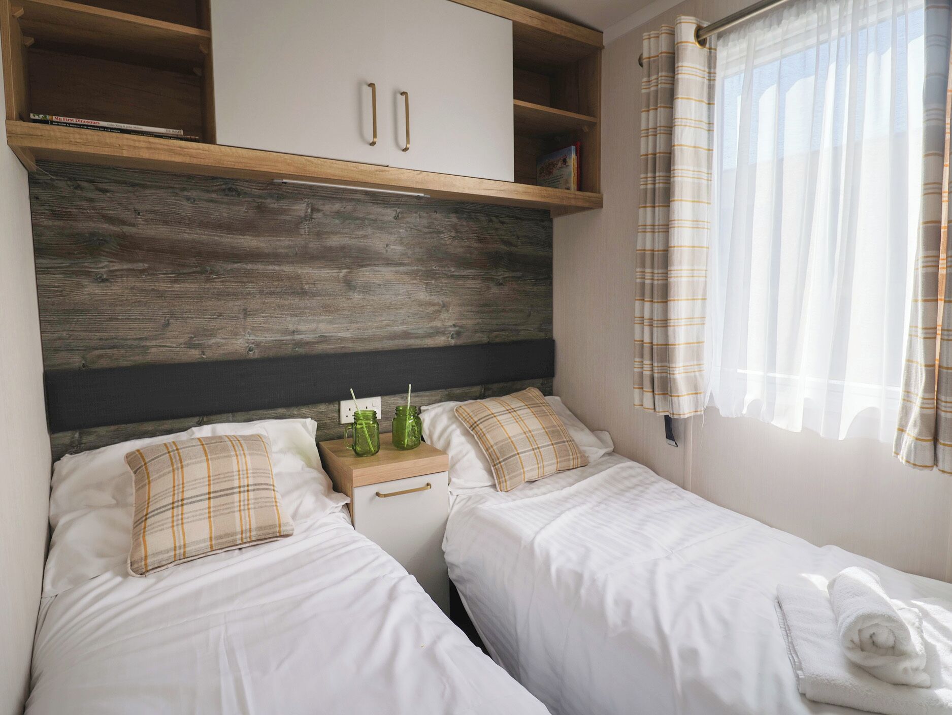 A bedroom with two beds, a wooden headboard, a nightstand with green glass jars, and a window with curtains.