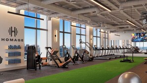 Ett gym med träningsutrustning, en vägg med varumärket ”HOMAGE”, stora fönster och utsikt över staden.